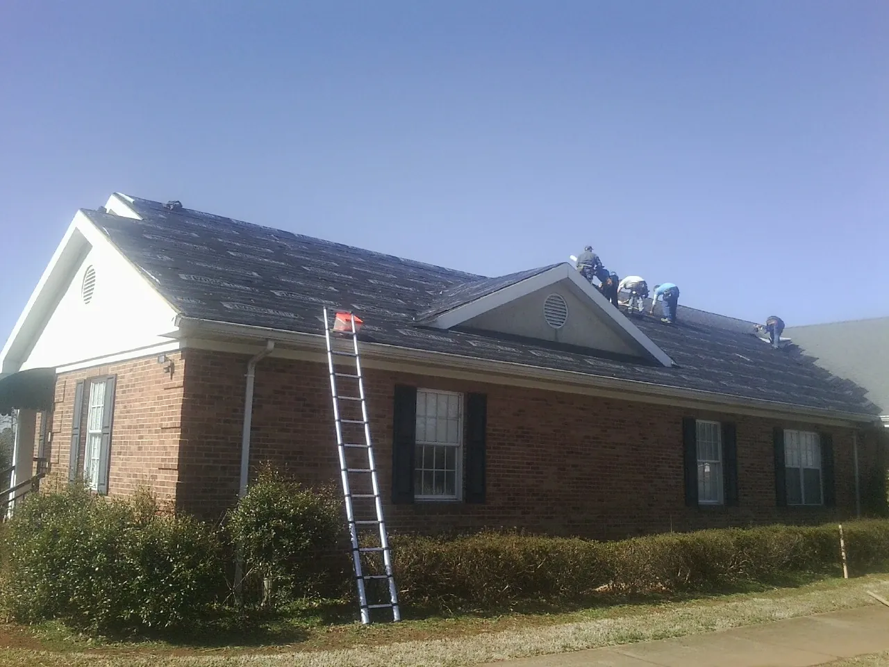 Crew tearing shingles off a brick ranch roof with a ladder set — Metal Roof Repair work by our crew in Wataga