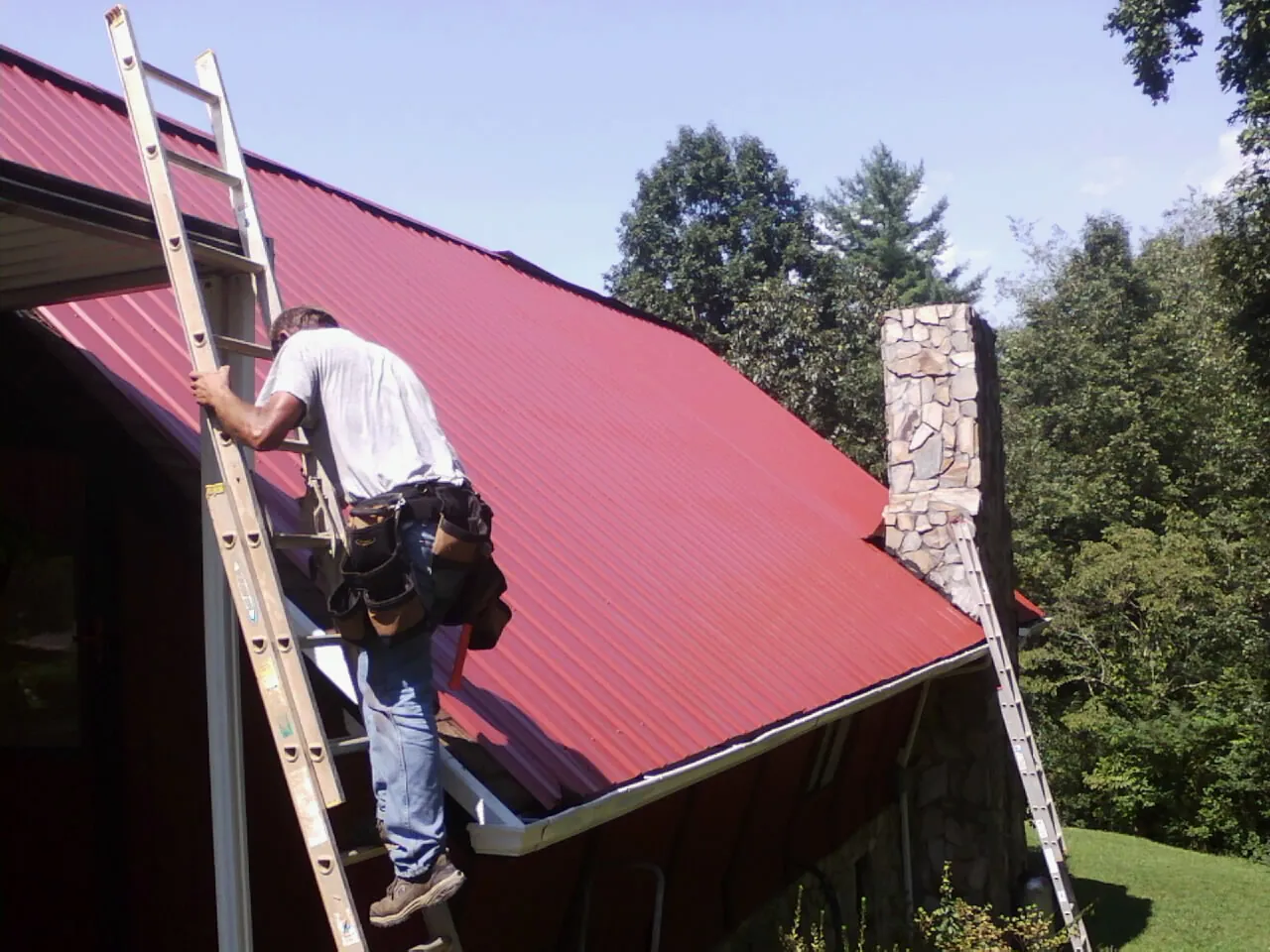 Roofer on a ladder beside a red metal roof and stone chimney — Soffit & Fascia Repair by Roofing Brothers in Wataga